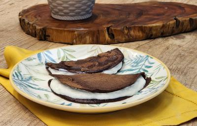 Fotografia em tons de marrom com um prato redondo ao centro. Em cima do prato existe duas panquecas de chocolate com aveia recheadas com um creme de iogurte.