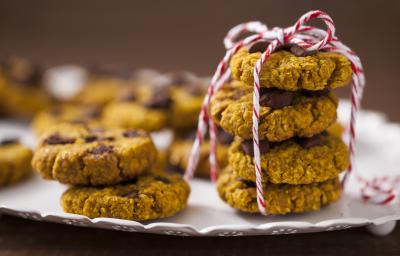 Fotografia em tons de amarelo em uma bancada de madeira com um recipiente branco raso e vários cookies integrais de aveia e abóbora em cima dele.