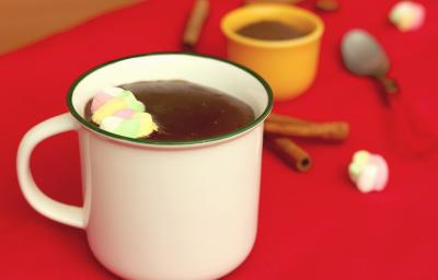 Fotografia em tons de branco e vermelho, ao centro uma xícara branca com chocolate quente e marshmallows, sobre toalha vermelha. Atrás um potinho com achocolatado e uma colher, com paus de canela e marshmallows espalhados ao redor.