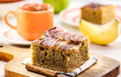 Fotografia de um pedaço de bolo de banana saudável, apoiado em um paninho branco, ao lado de dois paus de canela, sobre uma tábua pequena de madeira. Ao fundo tem uma caneca laranja e uma banana.