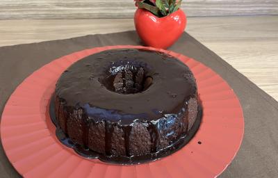 Fotografia de um bolo de chocolate e café com calda de chocolate sobre um apoio grande vermelho. O apoio está por cima de um pano marrom de tom escuro.