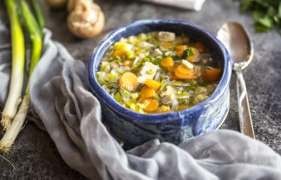 Fotografia em tons claros, no centro, um recipiente de vidro na cor azul. Dentro, uma sopa com cenoura, alho-poró e mandioquinha. Ao lado, uma colher de sopa, um alho-poró sobre a mesa. No fundo da foto, alguns champignons sobre a mesa.