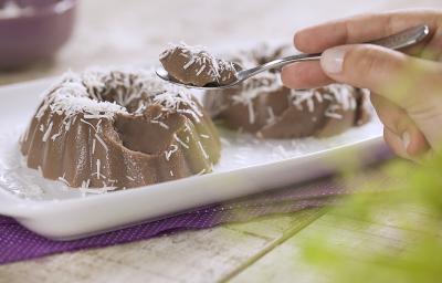 .Fotografia em tons de roxo e branco de uma bancada de madeira branca com um paninho roxo, sobre ele um prato retangular com dois gelados de dark chocolate. Sobre ele uma colher com um pedaço do gelado. Ao fundo um recipiente redondo com coco.