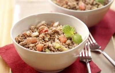 Fotografia em tons de vermelho em uma mesa de madeira clara com uma tábua de madeira, dois guardanapos de papel vermelhos e dois recipientes redondos com a salada de trigo com carne moída.