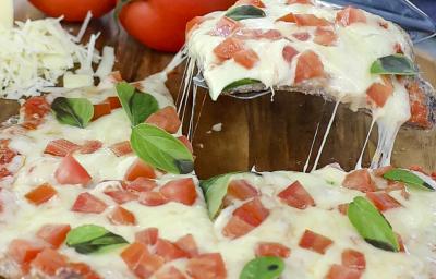 Foto aproximada de uma pizza marguerita, decorada com tomates e folhas de manjericão, sobre uma tábua de madeira e com tomates ao fundo