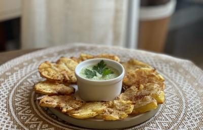 Fotografia em tons de marrom em uma bancada de madeira com uma toalha redonda branca de renda e um prato bege em cima com as batatas assadas e temperadas com um potinho branco ao centro com o molho de maionese verde.