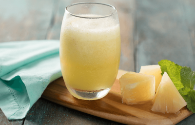 Fotografia em tons de verde em uma bancada de madeira mesclada, um pano verde água, uma tábua de madeira, um copo de vidro com a vitamina de abacaxi dentro, folhas de hortelã e pedaços de abacaxi ao lado.