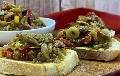 Foto da receita de caponata servida em duas fatias pão italiano e, atrás, em um bowl branco. Esses elementos estão sobre uma mesa de madeira redonda com um pano vermelho decorativo ao fundo