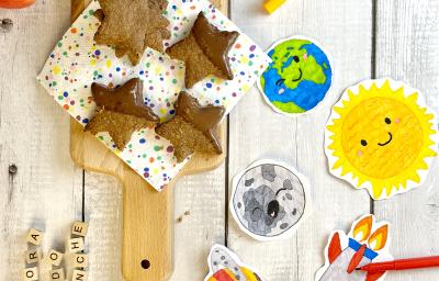 Fotografia em tons colorido em uma bancada de mandeira branca com uma tábua de madeira e as barrinhas de cereais em cima cobertas com metade de chocolate. Ao lado desenhos feitos com giz de cera.