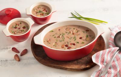 Fotografia em tons de branco e vermelho de uma bancada vista de cima. Em cima de uma tábua redonda contém um recipiente oval com alças nas cores vermelho e branco com a sopa e pedaços de pinhão por cima, ao lado dois potinhos com a sopa e um pano