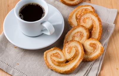 Foto da receita de Palmier ou Orelha de Macaco. Observa-se o biscoito em disposto em cima de um pano de prato e, ao lado, uma xícara com café sobre um pires.