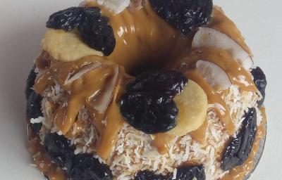 Foto da receita de Bolo de tapioca com doce de leite e ameixa. Observa-se um bolo coberto de doce de leite com cocos grudados na lateral e ameixas decorando.