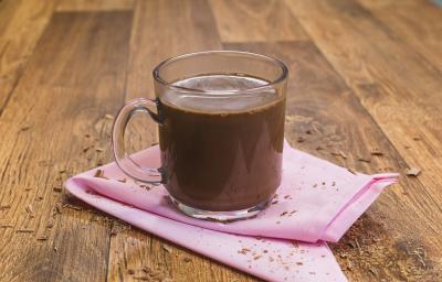 Fotografia de uma bancada de madeira com um paninho rosa e uma xícara transparente com o chocolate quente dentro.