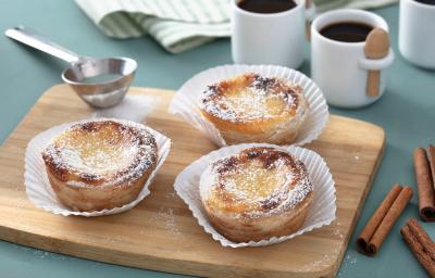 Fotografia em tons de amarelo em uma bancada de madeira de cor azul. Ao centro, uma tábua de madeira contendo os pasteis. Ao lado, há uma peneira de inox e ao fundo, duas xícaras de café.