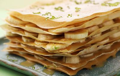 Fotografia em tons de verde em uma bancada de madeira com uma toalha verde, um prato quadrado com a torta crocante com recheio de doce de leite e bananas em rodelas.