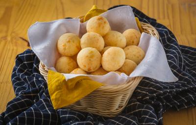 Foto aproximada de uma cesta de vime com diversos pães de queijo dentro. Ela está sobre uma bancada de madeira com um tecido azul quadriculado.