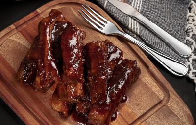 Foto de cima de uma tábua de madeira com a receita de costelinha suína ao molho barbecue, com alguns talheres de metal, um pote com mais molho e um tecido em tom cinza claro