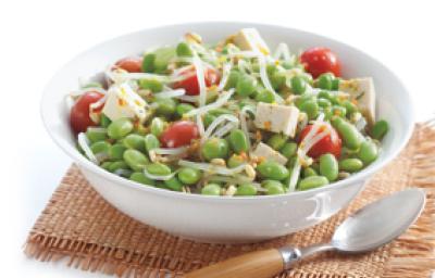 Fotografia em tons de marrom em um fundo branco com um paninho marrom e um prato fundo redondo branco com a salada de edamame, com soja verde, tomate e tofu.