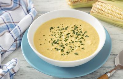 Fotografia em tons de amarelo e azul em uma bancada de madeira clara, um prato azul e branco ao centro com sopa de milho. Ao lado uma espiga de milho e um paninho azul quadriculado.