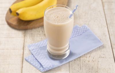 Foto em tons de bege com o copo de vitamina de banana com caramelo em cima de um paninho azul com bordas de bolinhas brancas, esses em cima de uma mesa de madeira. Fundo decorado com duas bananas nanicas em cima de uma tábua de madeira em formato oval.