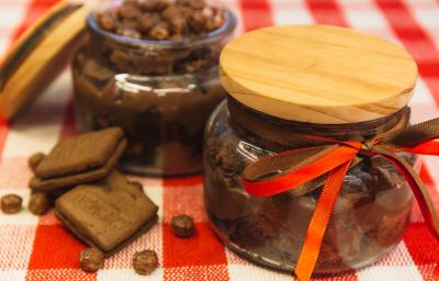 Foto de uma bancada forrada com uma toalha quadriculada vermelha e branca. Sobre ela há 2 potes de vidros com a receita de Brownie de Nescau com Brigadeiro. Um deles está fechado e o outro aberto. Além disso há 2 biscoitos e algumas bolinhas de cereal