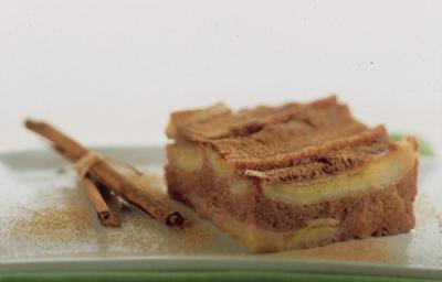 Fotografia em tons de canela e cinza, com pedaço de torta de banana com canela sobre travessa verde com ramos de canela ao lado, tudo polvilhado com canela, em fundo cinza.