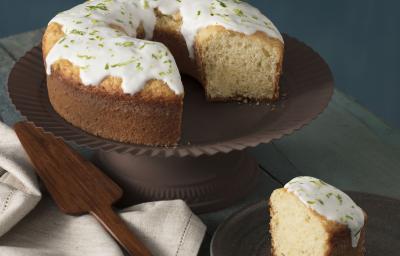 foto em tons de cinza, em uma mesa de madeira contém o bolo de iogurte ao lado um prato marrom com um pedaço de bolo um pano branco e por cima do pano uma espátula.