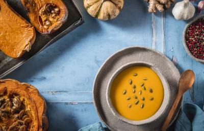 Fotografia vista de cima de um recipiente fundo de cerâmica com uma sopa de abóbora sobre um prato de cerâmica raso, com uma colher de madeira apoiada. Ao redor, várias abóboras assadas, pimentas em grãos e uma cabeça de alho, sobre uma mesa azul.