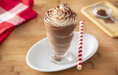 Foto da receita de Milkshake de Cappuccino. Observa-se um copo alto com a bebida decorado com chantilly e polvilhado de chocolate em pó. Ao lado direito, um canudo vermelho e branco para decorar.