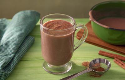 Fotografia em tons de marrom em uma bancada de madeira de cor verde. Ao centro, uma xícara contendo o chocolate quente. Ao lado, há uma toalha verde quadriculada e ao fundo, uma tábua de madeira com uma panela verde em cima.