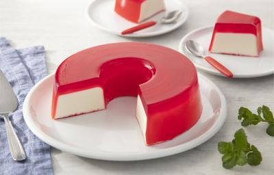 Fotografia em tons de rosa em uma bancada de madeira clara, um paninho azul, um prato branco redondo raso com a gelatina de morango recheada com creme branco. Ao fundo, pratinhos brancos com fatias da gelatina e talheres com cabo vermelho.