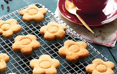 fotografia em tons de azul e bege de uma bancada azul vista de cima, contém uma grade com biscoitinhos em formato de flores e ao lado um pratinho vermelho com uma xicara vermelha que contém a bebida de chocolate branco.