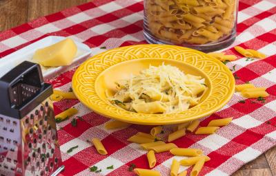 Foto de uma mesa com uma toalha xadrez vermelha e branca. Sobre ela há alguns macarrões crus, um ralador, um pedaço de queijo, um pote com mais macarrão e, ao centro, há um prato amarelo com a receita de Penne Alfredo.