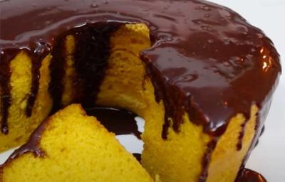 Foto da receita de Bolo de Cenoura com Cobertura de Chocolate. Observa-se um bolo com furo no meio bem de perto com uma fatia cortada e a cobertura de chocolate escorrendo