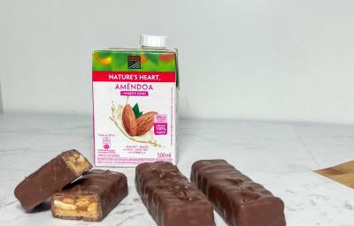 Foto em tons claros da receita de chocolate com caramelo e amendoim servida em 4 porções sobre uma mesa de mármore branca com uma embalagem de bebida vegetal de amêndoa nature's heart ao fundo