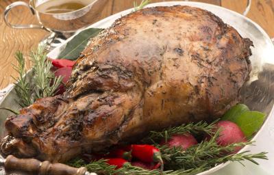 Fotografia em tons de marrom e verde, com travessa com duas alças contendo pernil assado guarnecido com ervas e pimenta vermelha, sobre dois guardanapos com um garfo, acima pratos empilhados, potinho e molheira com molhos, tudo sobre bancada de madeira.