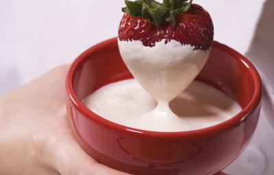 Fotografia de uma chef segurando um morango coberto de fondue de chocolate branco, que está dentro de um recipiente de vidro na cor vermelha.