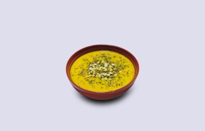 Fotografia em tons de amarelo em um fundo branco com um prato fundo vermelho e o caldo de abóbora e carne seca com Maggi dentro.