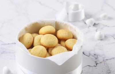 Fotografia em tons de branco de alguns biscoitos de massa branca dentro de uma cestinha branca com uma fita também branca amarrando-a. Ao fundo, alguns suspiros e um biscoito quebrado ao meio, sobre uma mesa de mármore branca.