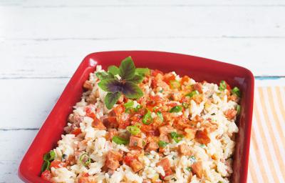 Fotografia em tons de vermelho e branco de uma bancada branca com uma pano listrado laranja e branco, uma travessa vermelha retangular com o arroz, pedaços de tomate e linguiça e manjericão.