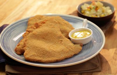 Fotografia em tons de marrom e azul de uma bancada de madeira clara, sobre ela um prato azul com dois bifes á milanesa e um mini recipiente refratário com molho.  Ao fundo um recipiente pequeno azul e redondo com vinagrete.