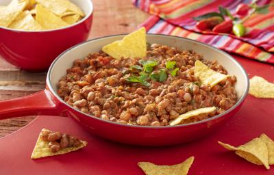 Fotografia em tons de vermelho em uma bancada de madeira escura, um jogo americano vermelho, um pano colorido, uma frigideira grande vermelho com o feijão mexicano dentro. Ao fundo, um pote vermelho com nachos.