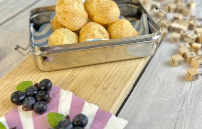 Fotografia em tons de roxo em uma bancada de madeira cinza com uma tábua de madeira marrom, uma lancheira em cima com pão de queijo feito de tapioca dentro dela. Ao lado, objetos e brinquedos infantis.