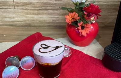 Fotografia em tons de vermelho em uma bancada de madeira, um pano vermelho e uma xícara com café e espuma rosa decorado com um coração com calda de chocolate ao centro. Ao fundo, um vaso vermelho com flores e em formato de coração.