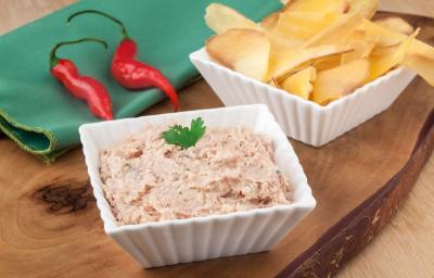 Fotografia em tons de branco, verde e marrom, com dois potinhos brancos contendo patê na cor rosa e mandioca chips, ao lado guardanapo verde com duas pimentas vermelhas, tudo sobre peça de madeira maciça.