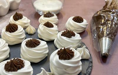 Foto da receita de vol au vent servida em diversas porções sobre uma base de porcelana  cinza escura em cima de uma mesa em tons de cor de rosa. Ao fundo há um copo de vidro com creme de leite e um saco com bico de confeitar com chocolate