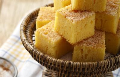 Foto de pedaços quadrados de bolo de fubá em um recipiente marrom em cima de um pano branco com detalhes laranja e azul