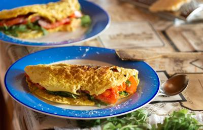 Fotografia em tons de azul e laranja em uma bancada de madeira escura, um prato redondo azul com uma omelete em cima dele recheada de tomate e rúcula. Ao lado colher e uns guardanapos marrons desenhados.