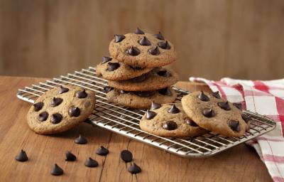 Fotografia em tons de marrom em uma bancada de madeira, um pano xadrez vermelho, um suporte de grelha com alguns cookies com gotas de chocolate em cima dele.