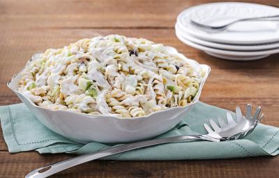 Fotografia em tons de azul em uma bancada de madeira escura, um pano azul com um recipiente redondo branco em cima dele e a salada de macarrão com peito de frango dentro dele. Ao lado, uma colher de servir macarrão.